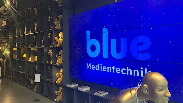 Ein blauer Bildschirm, auf dem „Blue Medientechnik“ steht, steht neben einem Regal mit goldenen Skulpturen. Ein Beispielbild für Orange Tontechnik München und Audiotechnik München.