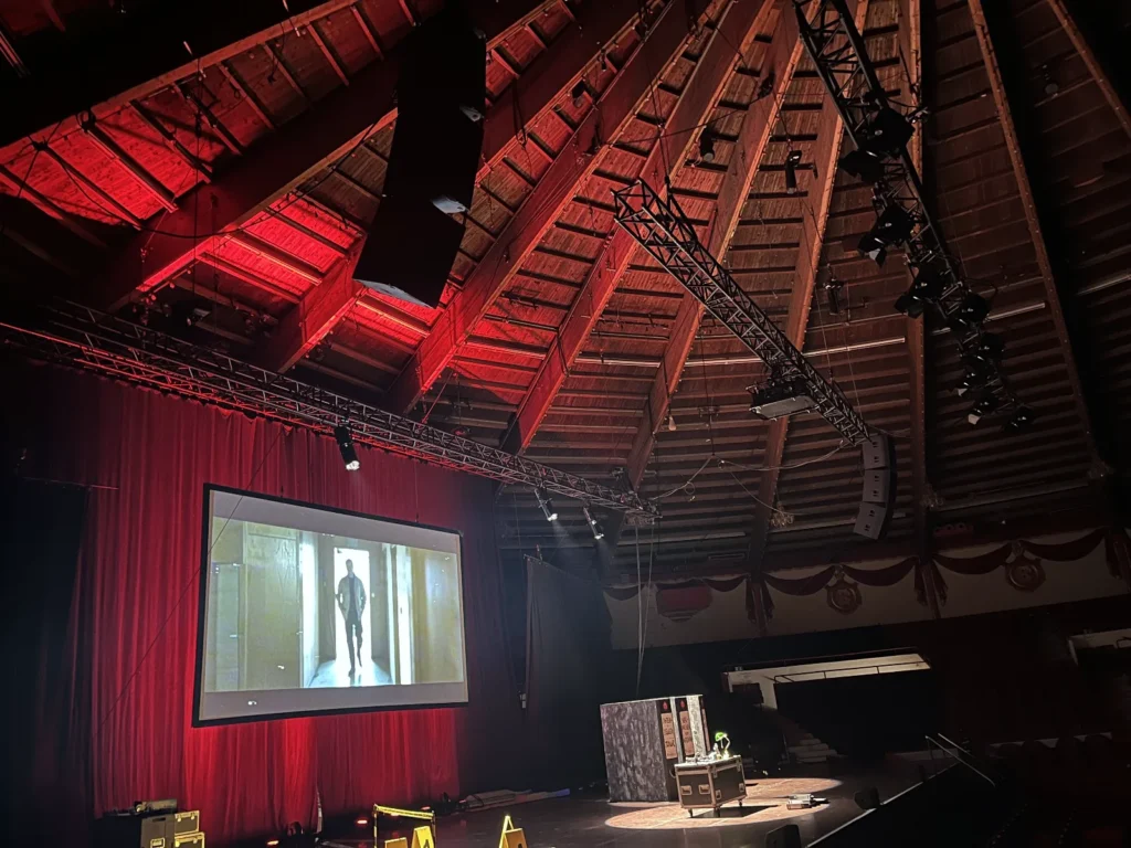 Innenansicht des Circus Krone mit Bühne, großer Projektionsfläche und Veranstaltungstechnik für Licht und Ton. Orange Veranstaltungstechnik Setup von Philipp Fleitner, inklusive Bühnenbeleuchtung und roter Beleuchtung der Holzdachkonstruktion. Totale der Tour-Produktion in der historischen Arena.